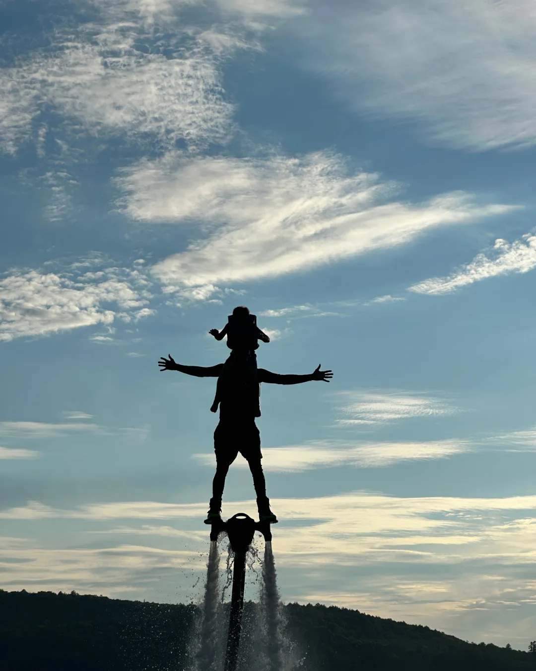 Adult standing on a Flyboard with a child seated on their shoulders against a cloudy Muskoka sky
